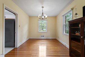 A small, empty dining room with light yellow walls, hardwood floors, a ceiling light fixture, two windows, and a glass-front cabinet in the corner. An open doorway leads to another room.