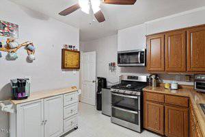 Kitchen with white cabinets, wooden countertops, brown upper cabinets, a stainless steel gas stove with oven, microwave, coffee maker, utensils, and a ceiling fan with light. White walls and neutral tile floor.