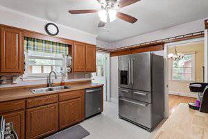 A kitchen with brown wooden cabinets, stainless steel appliances including a refrigerator, dishwasher, and sink, a ceiling fan with lights, and a window with a green plaid valance above the sink.