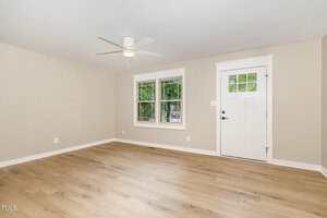 A bright, empty room with light wood flooring, beige walls, a white ceiling fan, a white door, and two windows showing green trees outside. White trim frames the windows, door, and baseboards.
