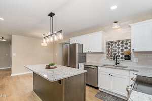A modern kitchen features white cabinets, stainless steel appliances, a light granite island with pendant lights above, wood flooring, and a decorative tile backsplash behind the sink.