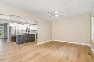 A spacious, empty room with light wood floors, beige walls, a white ceiling fan, and an open view of a modern kitchen with white cabinets, black island, hanging lights, and stainless steel appliances.