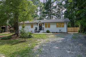 A single-story white house at 605 26th Street, Butner, with yellow-trimmed windows, a black front door, metal railings, and a gravel driveway, surrounded by trees and greenery.