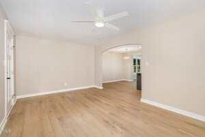 A spacious, empty room with light wood flooring, beige walls, a white ceiling fan, and an arched doorway leading to another room. Recessed lighting and baseboards are visible.