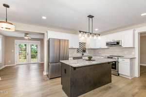 A modern kitchen with white cabinets, stainless steel appliances, a central island with granite countertop, pendant lighting, light wood flooring, and a view of a backyard through glass double doors.