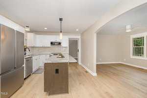 A modern kitchen with white cabinets, stainless steel appliances, an island with a dark base, pendant lighting, and light wood flooring, adjacent to an open living area with large windows.