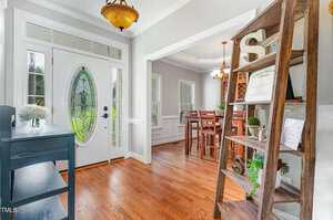 A bright entryway with a glass-paneled front door, wooden floors, a blue console table, and a tall wooden shelf. A dining room with a wooden table and chairs is visible through a large doorway.