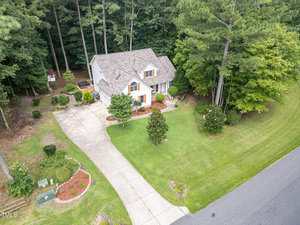 Aerial view of a two-story house with a light-colored exterior, a large driveway, landscaped front yard, and surrounded by tall trees and greenery along a street.