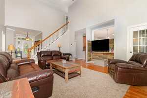 A spacious living room with brown leather sofas, a wooden coffee table, a beige rug, and a TV on a stand. A staircase with a wooden handrail leads upstairs, and hardwood floors are visible throughout.