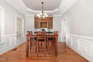 A dining room with hardwood floors, a wooden table and six chairs, a glass-front cabinet, large windows on the left, a chandelier overhead, and white paneled walls with crown molding.