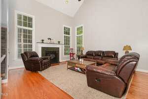 A living room with brown leather sofas, a matching recliner, a wooden coffee table on a beige rug, hardwood floors, a fireplace with a mantle, and three tall windows letting in natural light.