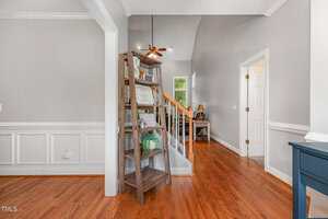 A wooden bookshelf stands near a staircase with wood steps and white railing in a home with light gray walls, white trim, and hardwood floors. A door leads to another room on the right.