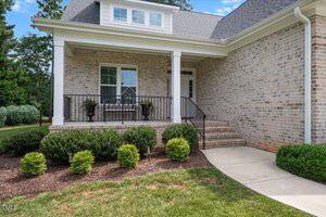 A brick house with a covered front porch, black metal railing, and steps leading up to the entrance. There are two potted plants by the window and neatly trimmed shrubs along the walkway.