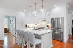 A modern white kitchen with an island, three upholstered barstools, stainless steel refrigerator, built-in oven, and pendant lights. Wood flooring adds warmth and cabinets line the walls.