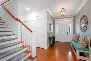 A bright entryway with hardwood floors, a staircase with white spindles and wood handrail, a white front door, a bench with cushions, a large wall clock, and a console table with flowers and decor.