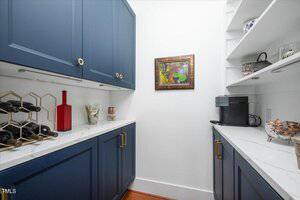 A small pantry with blue cabinets, white countertops, open shelves with dishes, a coffee maker, a wine rack with bottles, and a framed painting on a white wall.