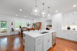 A modern kitchen with white cabinets, a large island with a sink, stainless steel appliances, pendant lights, and an adjacent dining area with wooden furniture and large windows overlooking greenery.