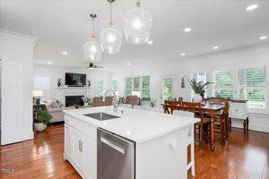 A modern open-concept kitchen and living area with white cabinetry, an island with a sink, stainless steel appliances, pendant lights, hardwood floors, and a dining table near large windows with plantation shutters.