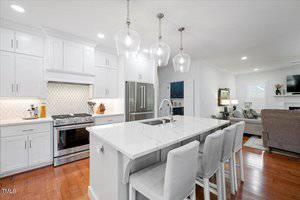 Modern white kitchen with stainless steel appliances, a large island with a built-in sink, three upholstered barstools, pendant lights overhead, and an open view into a living area with hardwood floors.