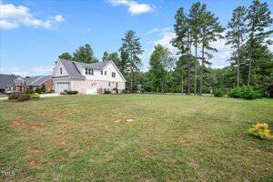A two-story brick and siding house with an attached garage sits on a spacious, well-maintained lawn with a few small shrubs, surrounded by tall trees under a partly cloudy sky.