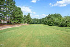 A neatly maintained golf fairway lined with tall pine trees on both sides under a partly cloudy sky; houses are partially visible through the trees on the left side.