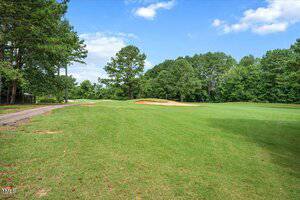 A golf course with green grass, a sand bunker in the distance, and trees lining the background under a partly cloudy sky. A paved path runs along the left side of the image.