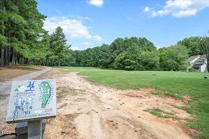 A golf course hole with a sign marking Hole 13, yardages, and a course map on the left; a dirt path runs alongside a grassy fairway bordered by trees, with a house visible in the distance on the right.
