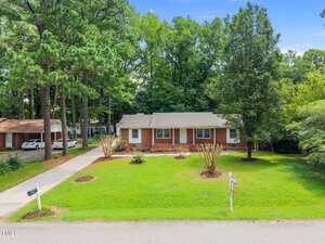 A single-story brick house with white trim sits on a well-maintained lawn with small landscaped areas and tall trees. A driveway runs along the left side, and a "for sale" sign is on the grass near the street.