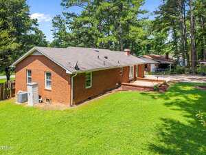 Single-story red brick house with a gray shingle roof, large grassy backyard, and a paved patio area, surrounded by tall trees and neighboring houses in the background.
