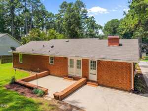 A single-story red brick house with a gray shingled roof. The back features double doors, a brick chimney, and a concrete patio area bordered by low brick walls, surrounded by grass and trees.