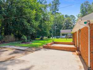 A concrete patio and driveway area beside a red brick house, bordered by a grassy lawn with landscaping and trees in the background. A wooden fence runs along the edge of the property.