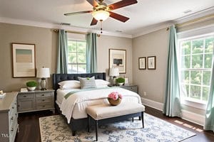 A bedroom with a large bed, gray tufted headboard, and white bedding. Two nightstands with lamps and decor flank the bed. Light blue curtains frame the windows, and a bench with a pink flower arrangement sits at the foot of the bed.