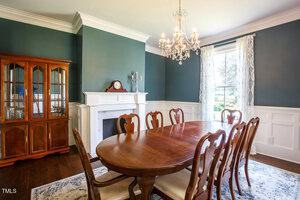 A dining room with a wooden table and eight chairs, a chandelier, a fireplace with a clock, a china cabinet, white wainscoting, dark green walls, hardwood floors, and a window with sheer curtains.