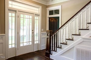 A bright entryway with a white front door featuring glass panels, sidelights, and a transom window. To the right, there is a staircase with white spindles, a dark wood handrail, and wood steps.