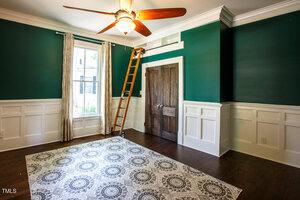 A room with dark green upper walls, white wainscoting, wooden floor, patterned area rug, double wooden doors, a window with beige curtains, and a wooden ladder leading to a loft storage area above the doors.