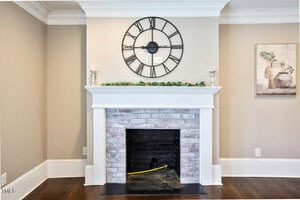 A brick fireplace with a white mantel is centered on a beige wall. A large black-framed analog clock hangs above the mantel, which is decorated with greenery and silver candle holders.
