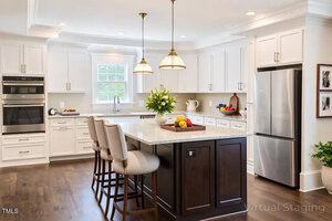 A modern kitchen with white cabinets, stainless steel appliances, and a dark wood island with three upholstered barstools. Two pendant lights hang above the island, which is decorated with flowers and bowls of fruit.