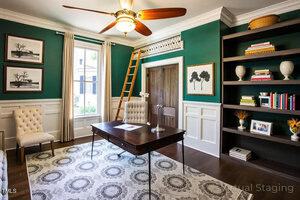 A home office with green walls, white wainscoting, a wooden desk, beige upholstered chairs, built-in shelves with books, framed art, a ceiling fan, a window with curtains, and a wooden ladder leading to a loft area.