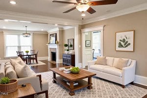 A bright living room with beige walls, a ceiling fan, a white sofa, a gray patterned rug, and a wooden coffee table. In the background is a dining area, fireplace, and large windows letting in natural light.