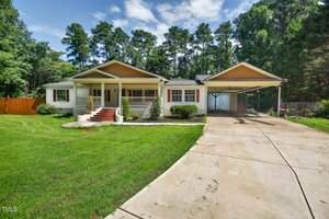 Charming single-story house at 3649 Windsor Drive, Franklinton, with white siding and yellow trim, a front porch with brick steps, attached carport, concrete driveway, green lawn, and trees under a partly cloudy sky.
