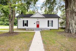 A white single-story house with black shutters and a red front door at 334 E Andrews Avenue in Henderson, set on a lawn with large trees and a concrete pathway leading to the entrance, photographed under a partly cloudy sky.
