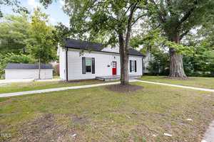 A small white house with black trim and a red front door sits on a corner lot with a grassy yard, concrete walkway, and mature trees. A detached white garage is visible in the background.