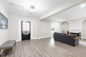 A spacious living room with light wood flooring, a black door, white walls, a dark sofa facing a white fireplace, a cushioned bench along one wall, and a framed painting of trees.