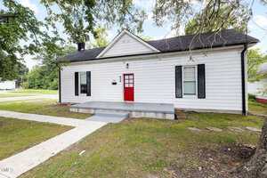 A single-story white house with black shutters, a red front door, and a small gray front porch. The yard has grass and trees, and a concrete walkway leads to the entrance.