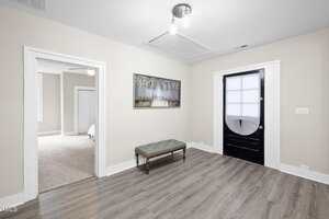 A small, empty room with light gray walls, wood flooring, a black door with a window shade, a wall bench, a framed painting, and an open doorway leading to a carpeted bedroom.