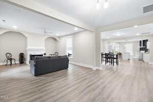 Spacious open-plan living area with light wood flooring, gray sofa, and fireplace, adjacent to a dining area and modern white kitchen with stainless steel appliances; neutral walls and bright lighting throughout.