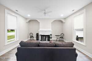 A living room with a dark sofa facing a white fireplace, two black metal bistro sets on each side, light wood floors, beige walls, and two large windows with white blinds.