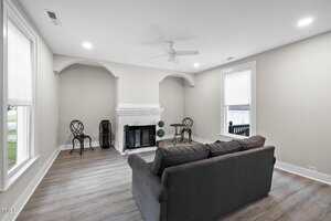 A living room with light gray walls, wood flooring, a dark gray sofa, a white fireplace, and two small round tables with chairs by the walls. Large windows bring in natural light.