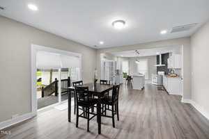 A dining area with a dark wood table and four chairs sits on wood flooring between a sliding glass door and an open kitchen with white cabinets, an island, and stainless steel appliances.