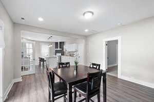 A modern dining area with a dark wood table and six chairs sits next to an open kitchen featuring white cabinets, a center island, and stainless steel appliances. The floor is wood and walls are light-colored.